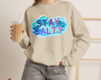 Puede incluir: Una sudadera beige de cuello redondo con un gráfico de olas oceánicas azules y el texto STAY SALTY impreso en letras moradas y rosas en el centro. La sudadera tiene mangas largas y acabados de canalé en el cuello, puños y dobladillo.