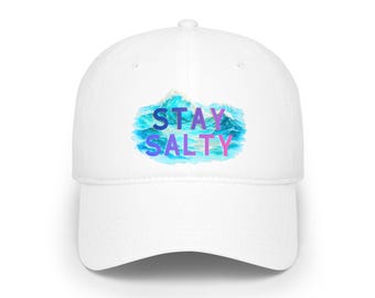 Puede incluir: Una gorra de béisbol blanca con un gráfico central de una ola oceánica azul. El texto STAY SALTY está impreso en una fuente con degradado púrpura y rosa en el centro de la ilustración de la ola, en el panel frontal de la gorra.