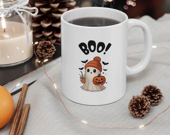 Puede incluir: Una taza de cerámica blanca con el gráfico de un fantasma que lleva un gorro naranja, sostiene una calabaza y una bebida, con el texto BOO! encima. Pequeños murciélagos negros y estrellas rodean al fantasma en el diseño.