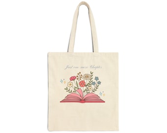 Puede incluir: Un bolso de tela color crema con un gráfico de un libro abierto de páginas rosas y un ramo de flores coloridas que emerge del centro. El texto "Just one more Chapter" está impreso sobre el libro, acompañado de pequeños iconos de estrellas.