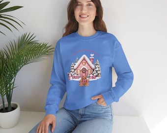 Puede incluir: Una sudadera azul brillante de manga larga con un gráfico central de una casa de jengibre rosa rodeada de pinos nevados, un muñeco de nieve y tres figuras de jengibre. El diseño incluye un pequeño corazón sobre la chimenea.