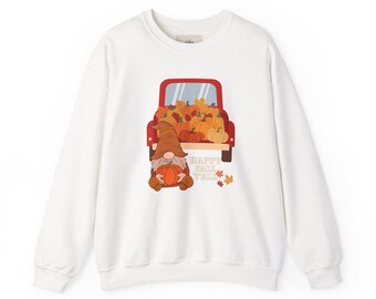 Puede incluir: Una sudadera blanca de cuello redondo con un gráfico de un camión rojo lleno de calabazas naranjas y hojas de otoño. Un gnomo con un sombrero marrón está sentado frente al camión sosteniendo una calabaza. El texto "HAPPY FALL Y'ALL" está impreso en marrón.