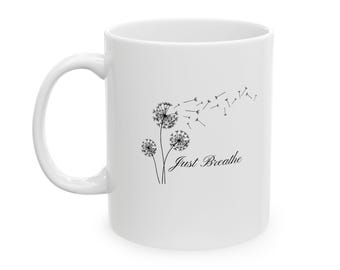 Puede incluir: Una taza de cerámica blanca con una ilustración de líneas negras de tres dientes de león con semillas volando con el viento. El texto Just Breathe está impreso en una fuente cursiva negra debajo de la ilustración floral.