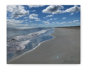 Puede incluir: Una fotografía de paisaje de una playa de arena que se encuentra con el océano bajo un cielo azul brillante con nubes blancas dispersas. Olas suaves llegan a la orilla reflejando la luz del sol. La firma Charmaine Hann es visible abajo a la derecha.