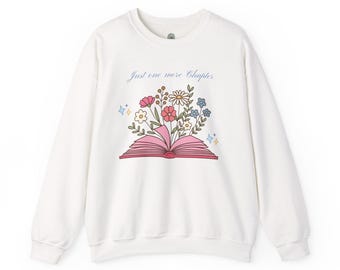 Puede incluir: Una sudadera blanca de manga larga con cuello redondo que presenta un gráfico de un libro abierto con flores coloridas saliendo de sus páginas. Sobre la ilustración, el texto Just one more Chapter aparece en azul con pequeñas estrellas.