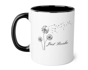 Puede incluir: Una taza de cerámica blanca con asa y borde interior negros. El exterior presenta una ilustración de líneas negras de tres dientes de león con semillas volando con el viento y el texto cursivo negro Just Breathe impreso debajo.