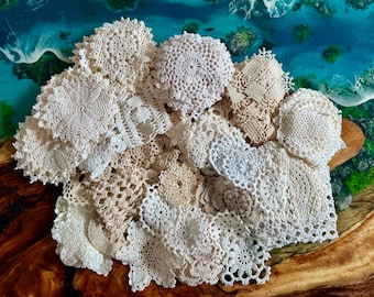May include: A collection of assorted round and square crochet doilies in various shades of white, cream, and beige. The pieces feature intricate openwork patterns, scalloped edges, and detailed threadwork, displayed in a dense, overlapping cluster.