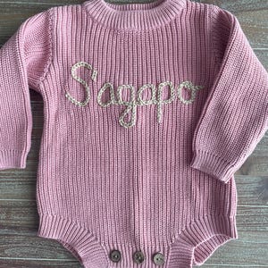 May include: A light pink knitted baby romper with long sleeves and a round neckline. The word "Sagapo" is embroidered in a cream-colored thread across the chest. Three wooden buttons are at the bottom. The romper is made of a soft, ribbed knit fabric.