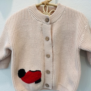 Peut inclure: Un cardigan en tricot rose clair avec un col rond et des boutons en bois. Le cardigan présente un motif appliqué rouge et noir sur le devant. Le vêtement est suspendu à un cintre en bois naturel.