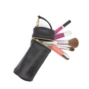 May include: Black cylindrical makeup bag with a gold zipper, handle, and hardware. The bag is open, revealing various makeup items, including lipsticks and brushes. The bag is made of textured material.