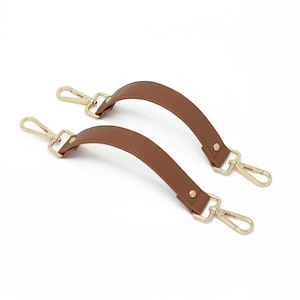 May include: Two brown leather bag handles with gold-colored metal clasps and hardware. The handles are curved and have a textured surface. The clasps are attached to the ends of the handles, ready to be attached to a bag.