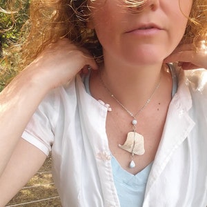 Könnte beinhalten: Eine Frau mit langen, lockigen, hellbraunen Haaren trägt ein weißes Hemd mit einem hellblauen Unterhemd. Sie trägt eine silberne Halskette mit einem weißen Muschelanhänger und einem hellblauen Stein.