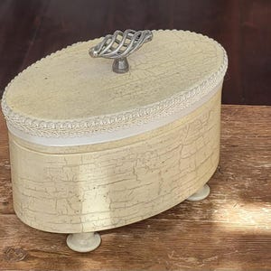 Może przedstawiać: Owalne, kremowe pudełko na drobiazgi z wykończeniem typu craquelé. Pokrywka posiada srebrny, skręcony uchwyt. Biała koronka otacza pokrywkę, a pudełko spoczywa na małych, zaokrąglonych nóżkach. Pudełko jest wystawione na drewnianej powierzchni.