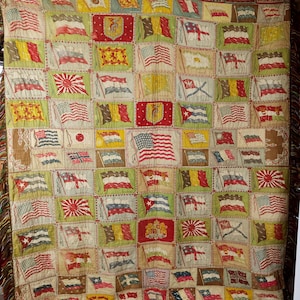 Puede incluir: Una manta textil tejida con un patrón de cuadrícula de varias banderas internacionales y escudos heráldicos en rojo, verde, amarillo y azul. El diseño incluye múltiples banderas estadounidenses y un borde con flecos en la parte inferior.