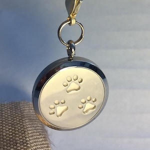 Puede incluir: Llavero de plata con un medallón redondo de plata con tres huellas de patas blancas. El medallón está unido a un cierre de resorte de plata y un anillo de plata.