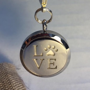 Puede incluir: Llavero de plata con un dije redondo difusor que dice "LOVE" con una huella de pata en el medio.
