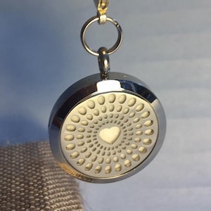 Puede incluir: Llavero de plata con un colgante redondo de difusor de plata. El difusor tiene un diseño en forma de corazón en el centro y un patrón de pequeños círculos alrededor del corazón.