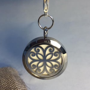 Puede incluir: Colgante difusor de aceites esenciales de aromaterapia plateado con un diseño floral decorativo. El colgante cuelga de un llavero y cierre plateados. El interior del colgante es de color crema claro.