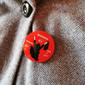 Peut inclure: Bouton rouge avec une silhouette noire d'une femme levant le poing et les mots "Femme", "Liberté", "Vie" en anglais et en farsi.