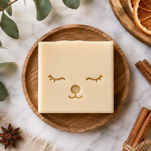 May include: A square, off-white bar of soap featuring an embossed, minimalist animal face design with closed eyes, a small circular nose, and a curved mouth. The soap is centered on a round, textured wooden plate with a visible wood grain pattern.