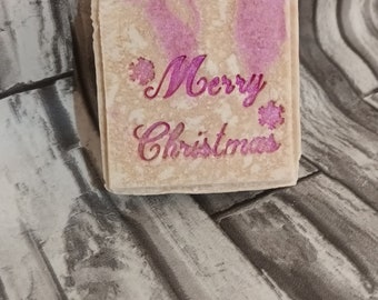 May include: A rectangular bar of soap with a light beige base and pink accents. The words "Merry Christmas" are written in pink script with snowflake accents. The soap has a textured appearance.