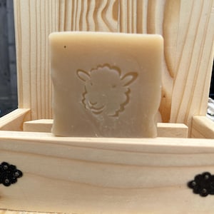 May include: A bar of natural soap in a wooden soap dish. The soap is light brown and has a sheep design embossed on it.