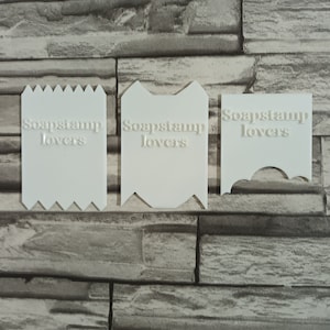 Può includere: Tre timbri per sapone in plastica bianca con il testo "Soapstamp lovers" su ciascuno. I timbri hanno forme e disegni diversi.