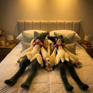 May include: Two plush toys featuring a character with a dark green pointed head, a pink collar, cream-coloured torso and arms, and long dark green legs. The plush figures have pointed ears, small facial features, and pink paw pads on their feet.