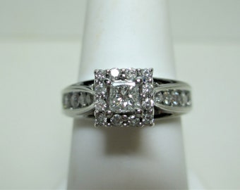 May include: A square-cut diamond ring with a halo of smaller diamonds. The ring features a band with additional diamond accents. The ring is set on a white background, showcasing its sparkle and design.
