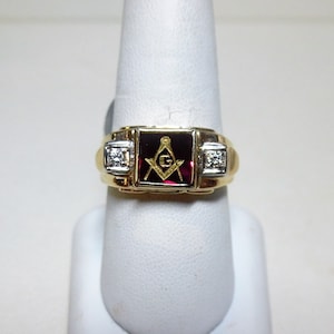 May include: A gold ring with a red stone and a gold Masonic symbol. The ring has two small diamonds on either side of the stone.
