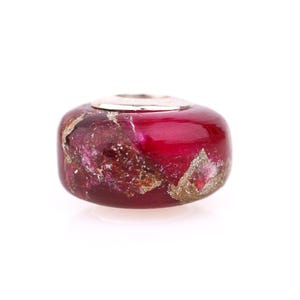 May include: A round, red glass bead with silver accents. The bead has a marbled appearance with gold and pink inclusions. The top of the bead has a silver-colored metal ring.