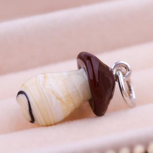 Op de afbeelding: Een paddenstoelvormige hanger met een cr&egrave;mekleurige steel en een donkerbruine dop. De steel heeft een zwart gebogen lijndetail. Een zilverkleurige ring is aan de dop bevestigd om op te hangen. De hanger is geplaatst tegen een zachte, roze achtergrond.