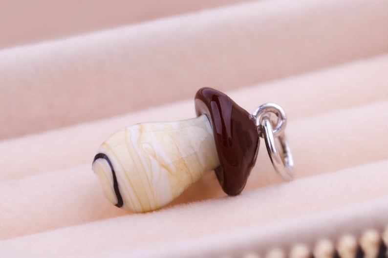 Op de afbeelding: Een paddenstoelvormige hanger met een cr&egrave;mekleurige steel en een donkerbruine dop. De steel heeft een zwart gebogen lijndetail. Een zilverkleurige ring is aan de dop bevestigd om op te hangen. De hanger is geplaatst tegen een zachte, roze achtergrond.