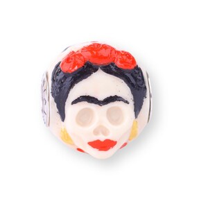May include: White ceramic bead featuring a painted image of a woman's face with a red flower crown. The woman's face is painted in a skull style with black hair and eyebrows, red lips, and yellow accents.