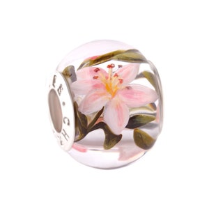 Puede incluir: Un abalorio de cristal transparente con una flor de lis rosa y hojas verdes en su interior. El abalorio tiene un anillo de metal plateado con las letras "GH" grabadas.
