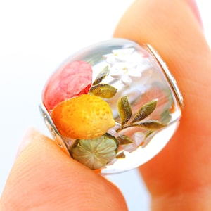 Könnte beinhalten: Ein transparenter Glasperlen-Anhänger mit einem bemalten Design aus einer Zitrone, einer rosa Blume und weißen Blumen mit grünen Blättern.