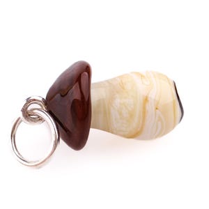 Op de afbeelding: Een paddenstoelvormige glazen hanger met een zilveren ring. De dop is donkerbruin van kleur en de steel is cr&egrave;mekleurig met een wervelpatroon. De hanger staat tegen een witte achtergrond.