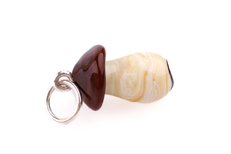Op de afbeelding: Een paddenstoelvormige glazen hanger met een zilveren ring. De dop is donkerbruin van kleur en de steel is cr&egrave;mekleurig met een wervelpatroon. De hanger staat tegen een witte achtergrond.