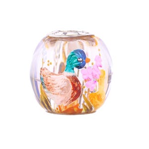 Può includere: Una perla di vetro rotonda e trasparente con una scena di anatra dipinta. L'anatra ha una testa blu, un collo verde e un corpo marrone. Fiori rosa e accenti gialli sono dipinti all'interno della perla.