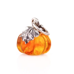 May include: A small, orange pumpkin charm with a silver-colored stem and leaf detail. The charm is likely made of glass or a similar material, and is set against a white background. A seasonal, fall-themed item.