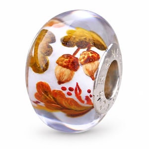 Pode incluir: Uma conta de vidro transparente com um núcleo branco apresentando motivos de outono pintados, incluindo bolotas castanhas, folhas de carvalho douradas e pequenas bagas vermelhas. A conta tem um núcleo de metal prateado com letras gravadas.