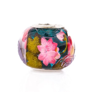 Può includere: Una perla rotonda di vetro con un motivo floreale. La perla presenta fiori rosa, foglie verdi e uno sfondo turchese. Ha un tappo in metallo color argento.