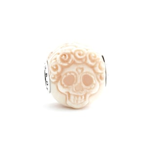 May include: White ceramic bead featuring a detailed skull design with floral accents. The skull has a heart-shaped nose and a floral crown.