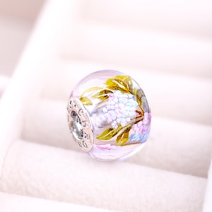 Peut inclure: Un charme de perle en verre transparent avec un motif floral. La perle est décorée de fleurs peintes à la main dans des tons de rose, violet et vert.