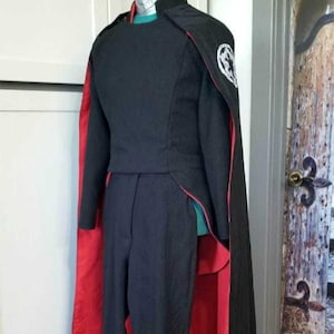 Könnte beinhalten: Ein schwarzes und rotes Kostüm mit einem Umhang und einem Symbol auf der Schulter. Das Kostüm ist aus einem Stoff, der wie ein Anzug aussieht.