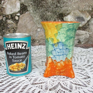 Puede incluir: Una lata azul y blanca de Heinz Baked Beans en salsa de tomate. La lata está sobre un encaje blanco de ganchillo. Un jarrón de cerámica colorido con un diseño azul, verde, amarillo y naranja está al lado de la lata.