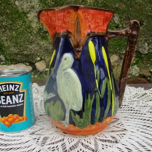 Może przedstawiać: Dzbanek ceramiczny w kolorze niebieskim i pomarańczowym z brązowym uchwytem. Dzbanek jest ozdobiony białym ptakiem i żółtymi kwiatami. Dzbanek stoi na białym obrusie z koronki.