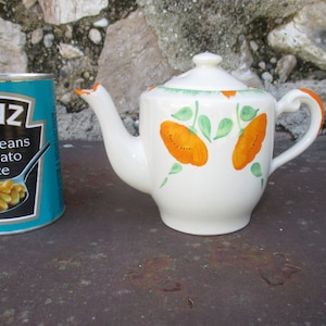 May include: A white teapot with an orange and green floral design. The teapot has a lid and a handle. The teapot is sitting on a brown surface.