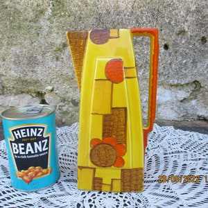 May include: A yellow ceramic pitcher with a geometric design in shades of orange, brown, and yellow. The pitcher has a handle and a spout. The pitcher is sitting on a white lace tablecloth.