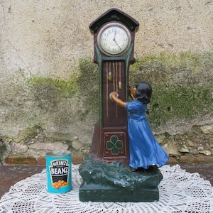 Puede incluir: Un reloj de pie vintage de color verde, marrón y rojo con una esfera blanca y agujas negras. Una figura de niña tira de las cadenas del reloj. El reloj está sentado sobre un encaje blanco.